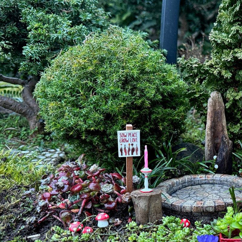 Miniature garden with a small sign, candles, and plants in a natural setting.